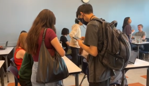 Students gather in a classroom setting; two students stand in the foreground—one with a large black bag and the other wearing a face mask and backpack, looking at a phone. Other students are seated at desks using laptops, with a clock on the wall showing the time.