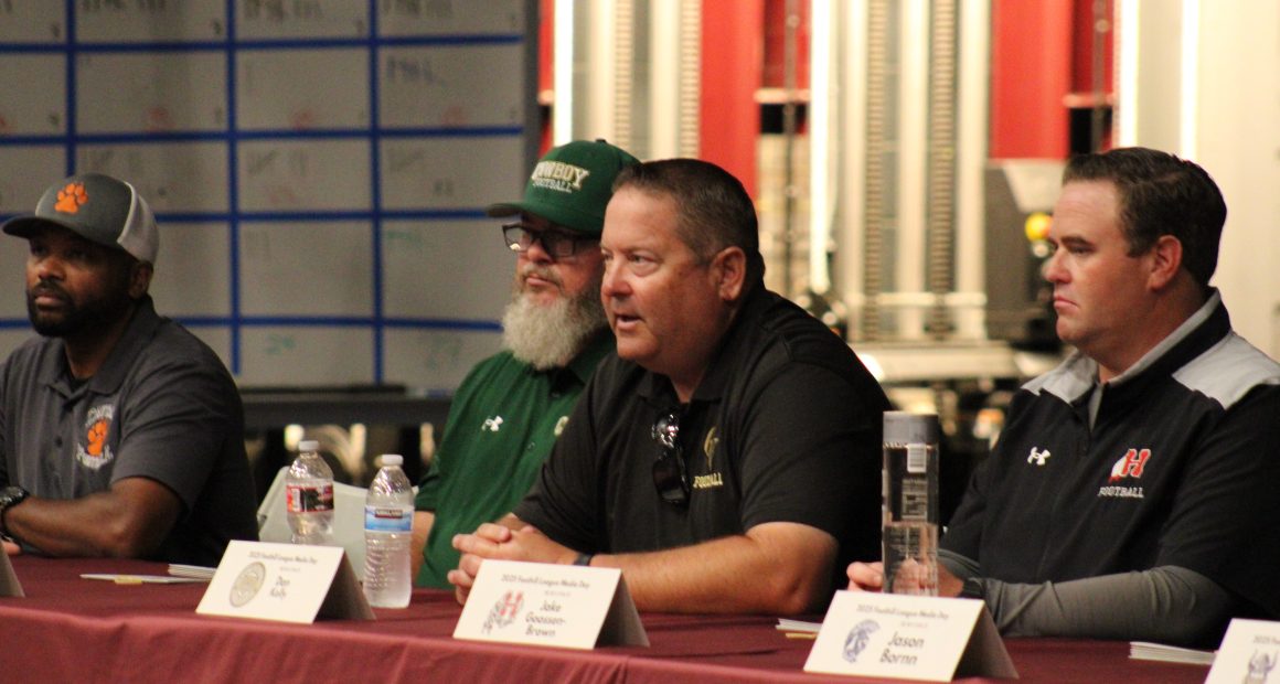Foothill High school football coaches Sirr Guy-Shakir,Ken Holsenbeck, Dan Kelly, Jake Goossen-Brown, and sit at a table during a Foothill League Media Day panel. Name placards, water bottles, and microphones are in front of them as they address the audience.