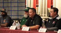 Foothill High school football coaches Sirr Guy-Shakir,Ken Holsenbeck, Dan Kelly, Jake Goossen-Brown, and sit at a table during a Foothill League Media Day panel. Name placards, water bottles, and microphones are in front of them as they address the audience.
