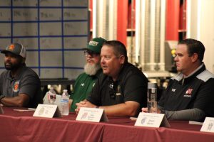 Foothill High school football coaches Sirr Guy-Shakir,Ken Holsenbeck, Dan Kelly, Jake Goossen-Brown, and sit at a table during a Foothill League Media Day panel. Name placards, water bottles, and microphones are in front of them as they address the audience.