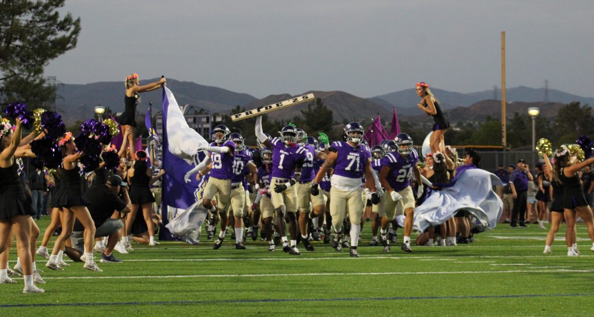 The Valencia Vikings football team in purple jerseys and tan pants runs onto the field through a banner, led by players holding flags and a sign reading “PRIDE 25.” at Valencia Field for the home opener vs The Simi Valley Pioneer's Cheerleaders dressed in black with colorful flower headbands cheer on both sides, some lifted into the air by teammates.