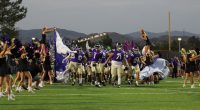The Valencia Vikings football team in purple jerseys and tan pants runs onto the field through a banner, led by players holding flags and a sign reading “PRIDE 25.” at Valencia Field for the home opener vs The Simi Valley Pioneer's Cheerleaders dressed in black with colorful flower headbands cheer on both sides, some lifted into the air by teammates.
