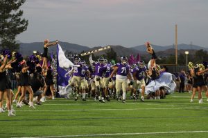 The Valencia Vikings football team in purple jerseys and tan pants runs onto the field through a banner, led by players holding flags and a sign reading “PRIDE 25.” at Valencia Field for the home opener vs The Simi Valley Pioneer's Cheerleaders dressed in black with colorful flower headbands cheer on both sides, some lifted into the air by teammates.