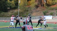 Hart quarterback Jacob in a gray and black uniform (number 2) preparing to throw the ball while being protected by his offensive line. Defensive players in white and red uniforms attempt to break through the blocks. A teammate wearing number 22 in gray is in the foreground, looking back toward the quarterback. The game is being played on a turf field with trees and a hillside in the background.