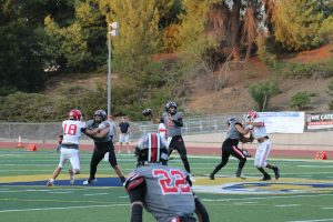 Hart quarterback Jacob in a gray and black uniform (number 2) preparing to throw the ball while being protected by his offensive line. Defensive players in white and red uniforms attempt to break through the blocks. A teammate wearing number 22 in gray is in the foreground, looking back toward the quarterback. The game is being played on a turf field with trees and a hillside in the background.