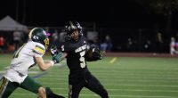 A Golden Valley High School football player in a black uniform (#5) carries the ball and stiff-arms an opposing defender in a white and green uniform (#11) during a night game on a turf field.