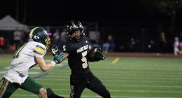 A Golden Valley High School football player in a black uniform (#5) carries the ball and stiff-arms an opposing defender in a white and green uniform (#11) during a night game on a turf field.
