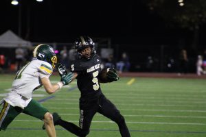 A Golden Valley High School football player in a black uniform (#5) carries the ball and stiff-arms an opposing defender in a white and green uniform (#11) during a night game on a turf field.