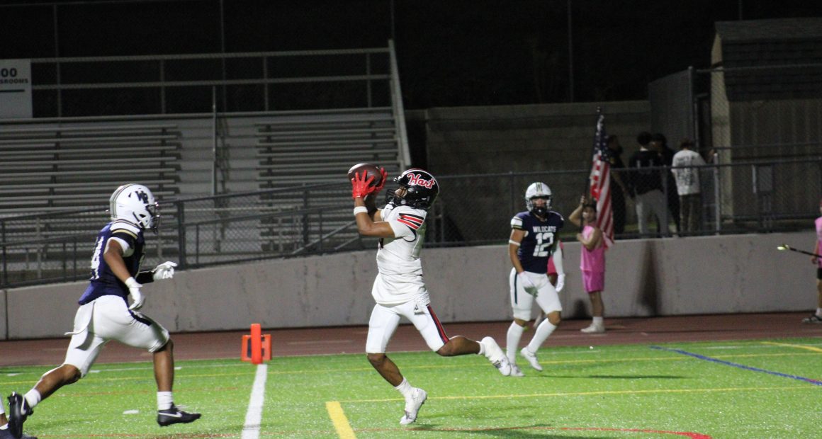 A Hart High School football player in a white uniform leaps to catch a pass during a night game against West Ranch, as a Wildcats defender closes in. The play takes place near the end zone with empty bleachers in the background.