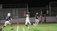 A Hart High School football player in a white uniform leaps to catch a pass during a night game against West Ranch, as a Wildcats defender closes in. The play takes place near the end zone with empty bleachers in the background.