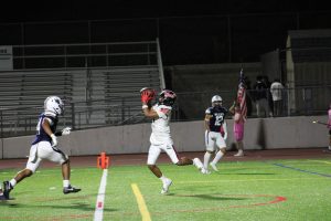 A Hart High School football player in a white uniform leaps to catch a pass during a night game against West Ranch, as a Wildcats defender closes in. The play takes place near the end zone with empty bleachers in the background.