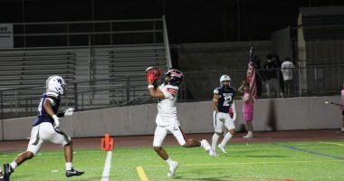 A Hart High School football player in a white uniform leaps to catch a pass during a night game against West Ranch, as a Wildcats defender closes in. The play takes place near the end zone with empty bleachers in the background.
