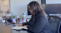 woman with long wavy brown hair wearing a black blazer is seated at a desk, writing in a notebook. She is facing slightly away from the camera. On the desk are colorful office supplies, a succulent plant, a water bottle with a logo, a nameplate, books, and computer monitors in the background.