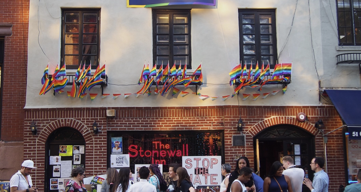 A crowd of people gathers outside The Stonewall Inn, a historic LGBTQ+ landmark in New York City. The building is decorated with numerous rainbow Pride flags and a banner above the windows that reads "#FindYourPark." Signs displayed include messages like "STOP THE HATE" and "LOVE > HATE." The scene reflects support for LGBTQ+ rights and commemorates the legacy of Stone