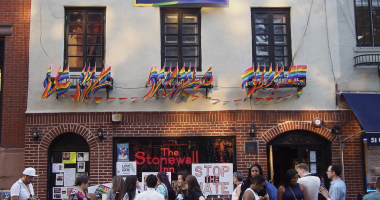 A crowd of people gathers outside The Stonewall Inn, a historic LGBTQ+ landmark in New York City. The building is decorated with numerous rainbow Pride flags and a banner above the windows that reads "#FindYourPark." Signs displayed include messages like "STOP THE HATE" and "LOVE > HATE." The scene reflects support for LGBTQ+ rights and commemorates the legacy of Stone