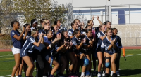 A girls' flag football team poses for a group photo on a turf field. They are wearing navy blue and white jerseys, some with knee braces and flags. The players smile and make hand signs, with a school building and fence in the background.