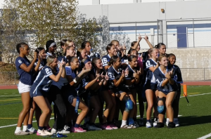 A girls' flag football team poses for a group photo on a turf field. They are wearing navy blue and white jerseys, some with knee braces and flags. The players smile and make hand signs, with a school building and fence in the background.