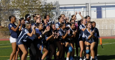 A girls' flag football team poses for a group photo on a turf field. They are wearing navy blue and white jerseys, some with knee braces and flags. The players smile and make hand signs, with a school building and fence in the background.