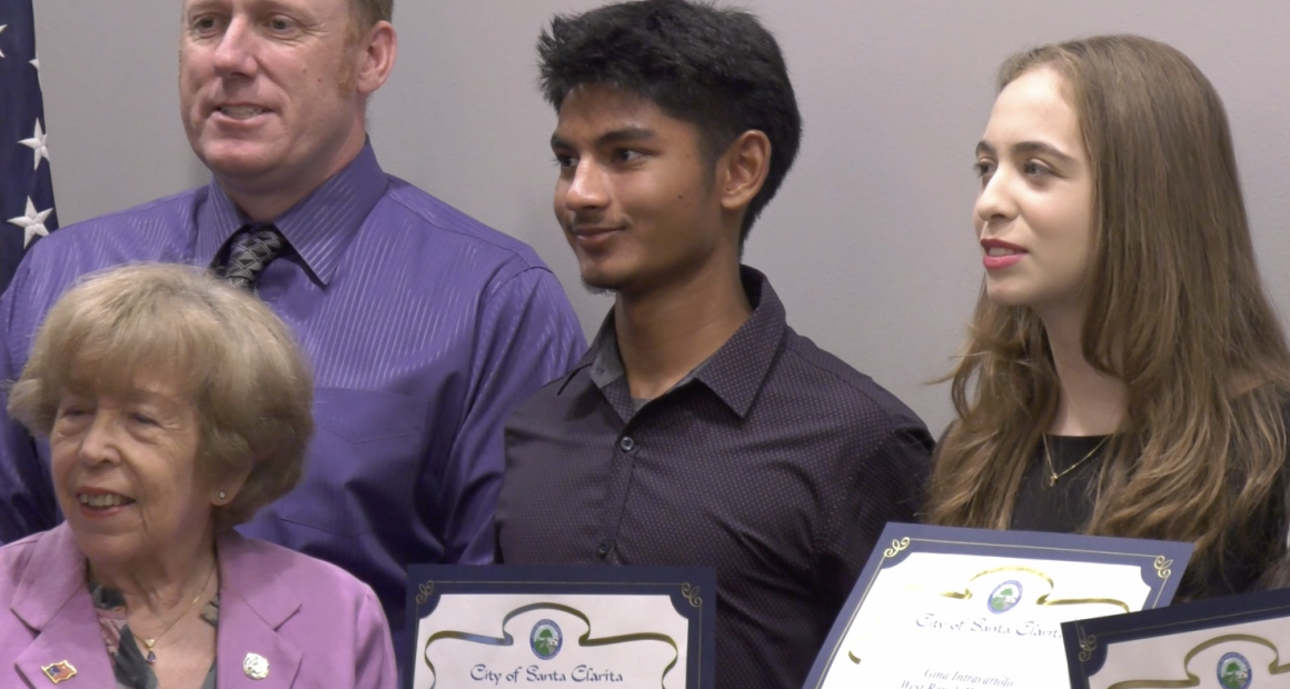 A group of five people pose for a photo, with three young individuals holding certificates from the City of Santa Clarita. The certificates recognize high school achievements. The group includes an older woman in a lavender blazer, a man in a purple shirt and tie, and three students standing in the center and right, smiling and dressed in formal or semi-formal attire. An American flag is partially visible in the background.
