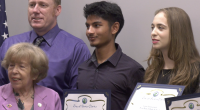 A group of five people pose for a photo, with three young individuals holding certificates from the City of Santa Clarita. The certificates recognize high school achievements. The group includes an older woman in a lavender blazer, a man in a purple shirt and tie, and three students standing in the center and right, smiling and dressed in formal or semi-formal attire. An American flag is partially visible in the background.