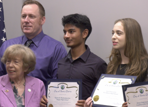 A group of five people pose for a photo, with three young individuals holding certificates from the City of Santa Clarita. The certificates recognize high school achievements. The group includes an older woman in a lavender blazer, a man in a purple shirt and tie, and three students standing in the center and right, smiling and dressed in formal or semi-formal attire. An American flag is partially visible in the background.
