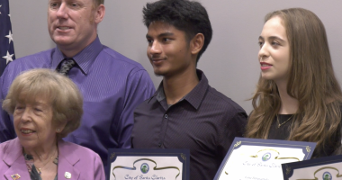 A group of five people pose for a photo, with three young individuals holding certificates from the City of Santa Clarita. The certificates recognize high school achievements. The group includes an older woman in a lavender blazer, a man in a purple shirt and tie, and three students standing in the center and right, smiling and dressed in formal or semi-formal attire. An American flag is partially visible in the background.