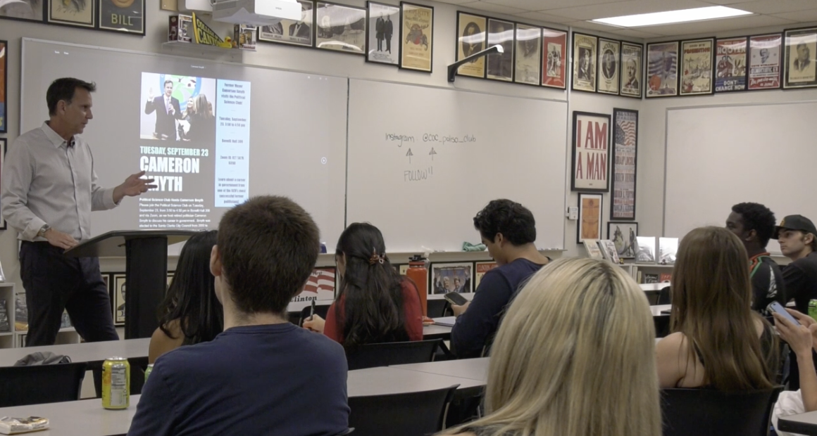 A man stands at a podium giving a presentation to a classroom of college students. A slide on the screen behind him promotes an event featuring Cameron Smyth on Tuesday, September 23. The students are seated at desks, some taking notes or using their phones. The classroom walls are decorated with historical and political posters, and a whiteboard behind the speaker displays the club's Instagram handle "@coc_polsa_club" with the word "FOLLOW" written below.