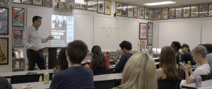 A man stands at a podium giving a presentation to a classroom of college students. A slide on the screen behind him promotes an event featuring Cameron Smyth on Tuesday, September 23. The students are seated at desks, some taking notes or using their phones. The classroom walls are decorated with historical and political posters, and a whiteboard behind the speaker displays the club's Instagram handle "@coc_polsa_club" with the word "FOLLOW" written below.