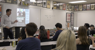A man stands at a podium giving a presentation to a classroom of college students. A slide on the screen behind him promotes an event featuring Cameron Smyth on Tuesday, September 23. The students are seated at desks, some taking notes or using their phones. The classroom walls are decorated with historical and political posters, and a whiteboard behind the speaker displays the club's Instagram handle "@coc_polsa_club" with the word "FOLLOW" written below.