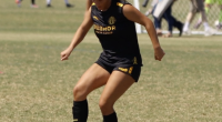 A soccer player in a black uniform with yellow lettering and accents dribbles the ball on a grass field during a match, with the goal and other players visible in the background.