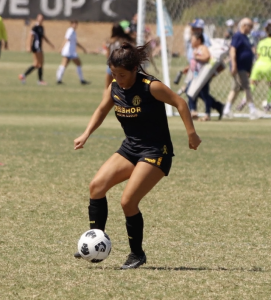 A soccer player in a black uniform with yellow lettering and accents dribbles the ball on a grass field during a match, with the goal and other players visible in the background.