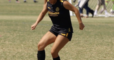 A soccer player in a black uniform with yellow lettering and accents dribbles the ball on a grass field during a match, with the goal and other players visible in the background.