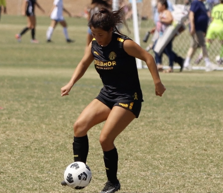 A soccer player in a black uniform with yellow lettering and accents dribbles the ball on a grass field during a match, with the goal and other players visible in the background.