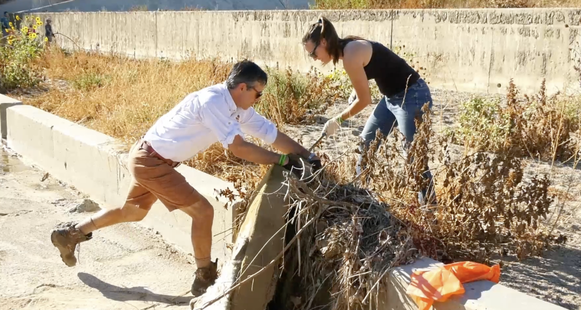 Two people are cleaning debris from a dry, overgrown channel. A man in shorts and boots pushes against a concrete edge while pulling at vegetation, and a woman in jeans and gloves helps remove tangled plants.