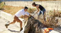 Two people are cleaning debris from a dry, overgrown channel. A man in shorts and boots pushes against a concrete edge while pulling at vegetation, and a woman in jeans and gloves helps remove tangled plants.