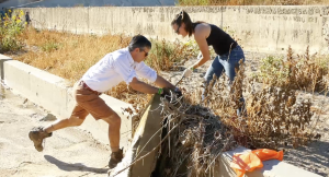 Two people are cleaning debris from a dry, overgrown channel. A man in shorts and boots pushes against a concrete edge while pulling at vegetation, and a woman in jeans and gloves helps remove tangled plants.