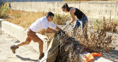 Two people are cleaning debris from a dry, overgrown channel. A man in shorts and boots pushes against a concrete edge while pulling at vegetation, and a woman in jeans and gloves helps remove tangled plants.