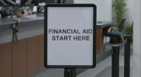 A sign in a financial aid office at College of the Canyons reads “FINANCIAL AID START HERE,” placed at the beginning of a line with desks and chairs visible in the background.