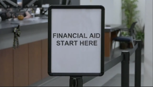 A sign in a financial aid office at College of the Canyons reads “FINANCIAL AID START HERE,” placed at the beginning of a line with desks and chairs visible in the background.