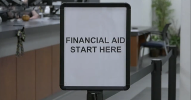 A sign in a financial aid office at College of the Canyons reads “FINANCIAL AID START HERE,” placed at the beginning of a line with desks and chairs visible in the background.