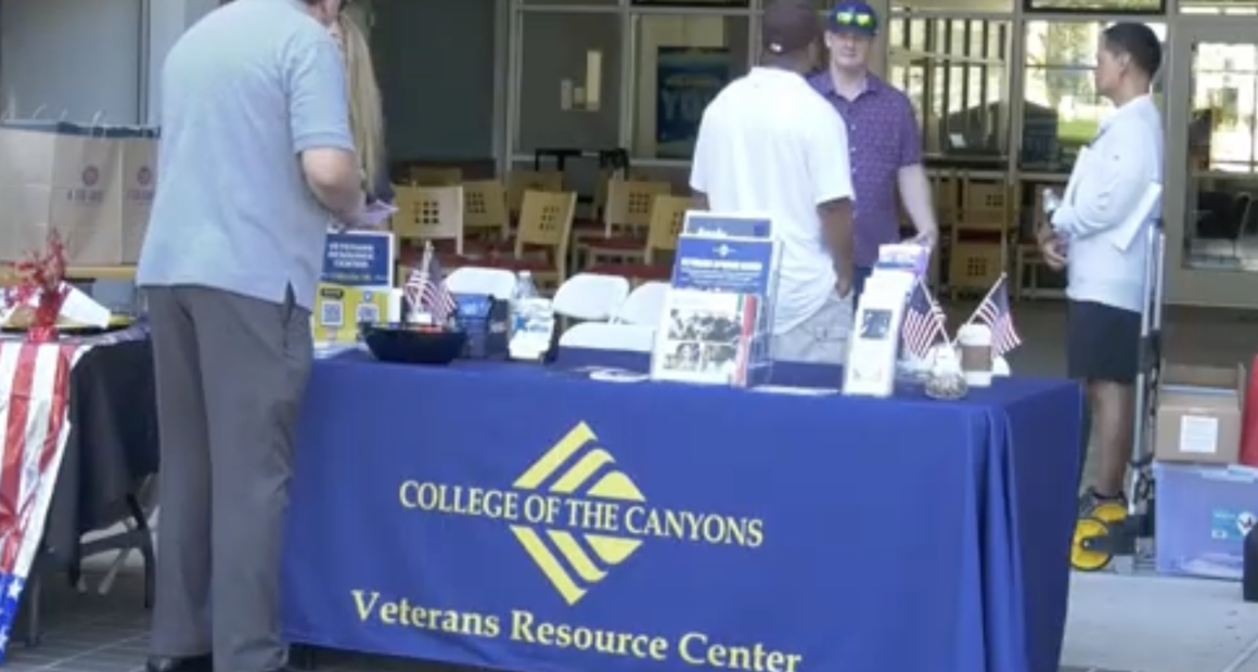 A table with a blue tablecloth reading “College of the Canyons Veterans Resource Center” is set up outdoors, displaying brochures, small American flags, and informational materials. Several people stand around the table engaged in conversation. In the background, chairs and tables are visible inside a building.