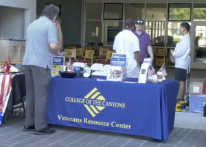 A table with a blue tablecloth reading “College of the Canyons Veterans Resource Center” is set up outdoors, displaying brochures, small American flags, and informational materials. Several people stand around the table engaged in conversation. In the background, chairs and tables are visible inside a building.
