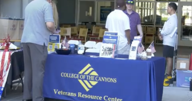 A table with a blue tablecloth reading “College of the Canyons Veterans Resource Center” is set up outdoors, displaying brochures, small American flags, and informational materials. Several people stand around the table engaged in conversation. In the background, chairs and tables are visible inside a building.
