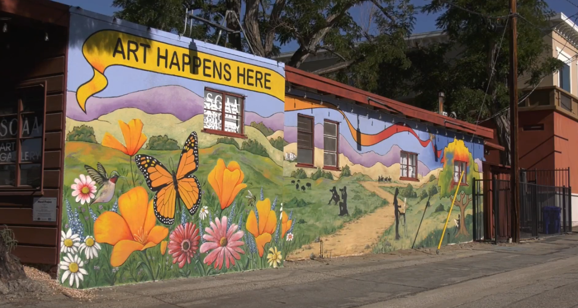 A colorful mural painted on the side of a building features bright orange poppies, pink and white daisies, a hummingbird, and a large monarch butterfly in the foreground. Behind them, rolling green hills and purple mountains stretch into the distance. Silhouettes of people and a path through the landscape are also depicted. Above the mural, a yellow banner reads “ART HAPPENS HERE.” The building appears to be an art gallery, with windows and a sign visible on the side.