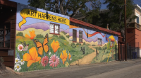 A colorful mural painted on the side of a building features bright orange poppies, pink and white daisies, a hummingbird, and a large monarch butterfly in the foreground. Behind them, rolling green hills and purple mountains stretch into the distance. Silhouettes of people and a path through the landscape are also depicted. Above the mural, a yellow banner reads “ART HAPPENS HERE.” The building appears to be an art gallery, with windows and a sign visible on the side.