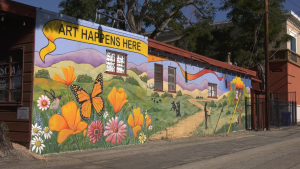 A colorful mural painted on the side of a building features bright orange poppies, pink and white daisies, a hummingbird, and a large monarch butterfly in the foreground. Behind them, rolling green hills and purple mountains stretch into the distance. Silhouettes of people and a path through the landscape are also depicted. Above the mural, a yellow banner reads “ART HAPPENS HERE.” The building appears to be an art gallery, with windows and a sign visible on the side.