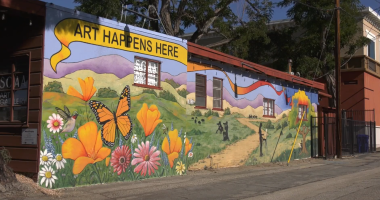 A colorful mural painted on the side of a building features bright orange poppies, pink and white daisies, a hummingbird, and a large monarch butterfly in the foreground. Behind them, rolling green hills and purple mountains stretch into the distance. Silhouettes of people and a path through the landscape are also depicted. Above the mural, a yellow banner reads “ART HAPPENS HERE.” The building appears to be an art gallery, with windows and a sign visible on the side.