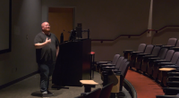 MAX KELLER ASSISTANT PROFESSOR AND CHAIR OF THE CINEMA DEPARTMENT stands at the front of a dimly lit lecture hall or auditorium. He is positioned next to a dark-colored podium with a monitor, and he is gesturing with his right hand as if speaking or presenting. The hall has rows of empty, reddish-brown upholstered seats that curve around the front stage area. The floor is carpeted in red. A door is visible behind him to the left of the podium.