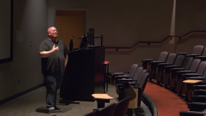 MAX KELLER ASSISTANT PROFESSOR AND CHAIR OF THE CINEMA DEPARTMENT stands at the front of a dimly lit lecture hall or auditorium. He is positioned next to a dark-colored podium with a monitor, and he is gesturing with his right hand as if speaking or presenting. The hall has rows of empty, reddish-brown upholstered seats that curve around the front stage area. The floor is carpeted in red. A door is visible behind him to the left of the podium.