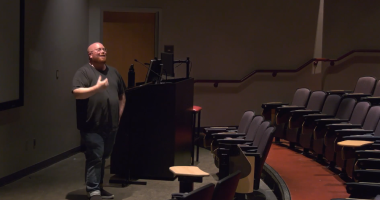 MAX KELLER ASSISTANT PROFESSOR AND CHAIR OF THE CINEMA DEPARTMENT stands at the front of a dimly lit lecture hall or auditorium. He is positioned next to a dark-colored podium with a monitor, and he is gesturing with his right hand as if speaking or presenting. The hall has rows of empty, reddish-brown upholstered seats that curve around the front stage area. The floor is carpeted in red. A door is visible behind him to the left of the podium.